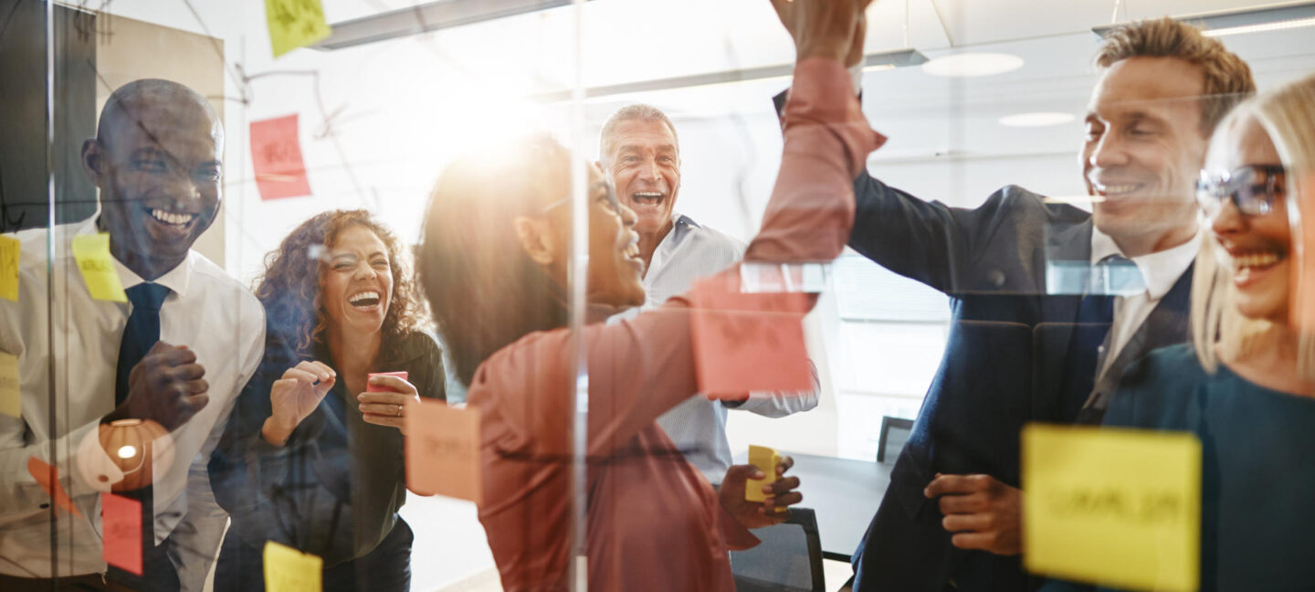Two diverse businesspeople high fiving together after a brainstorming session in an office with laughing colleagues looking on