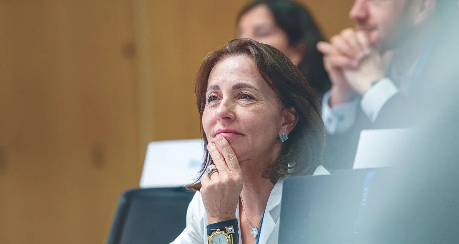 A woman attentively listening during an executive education session, with other participants blurred in the background. - IMD Business School