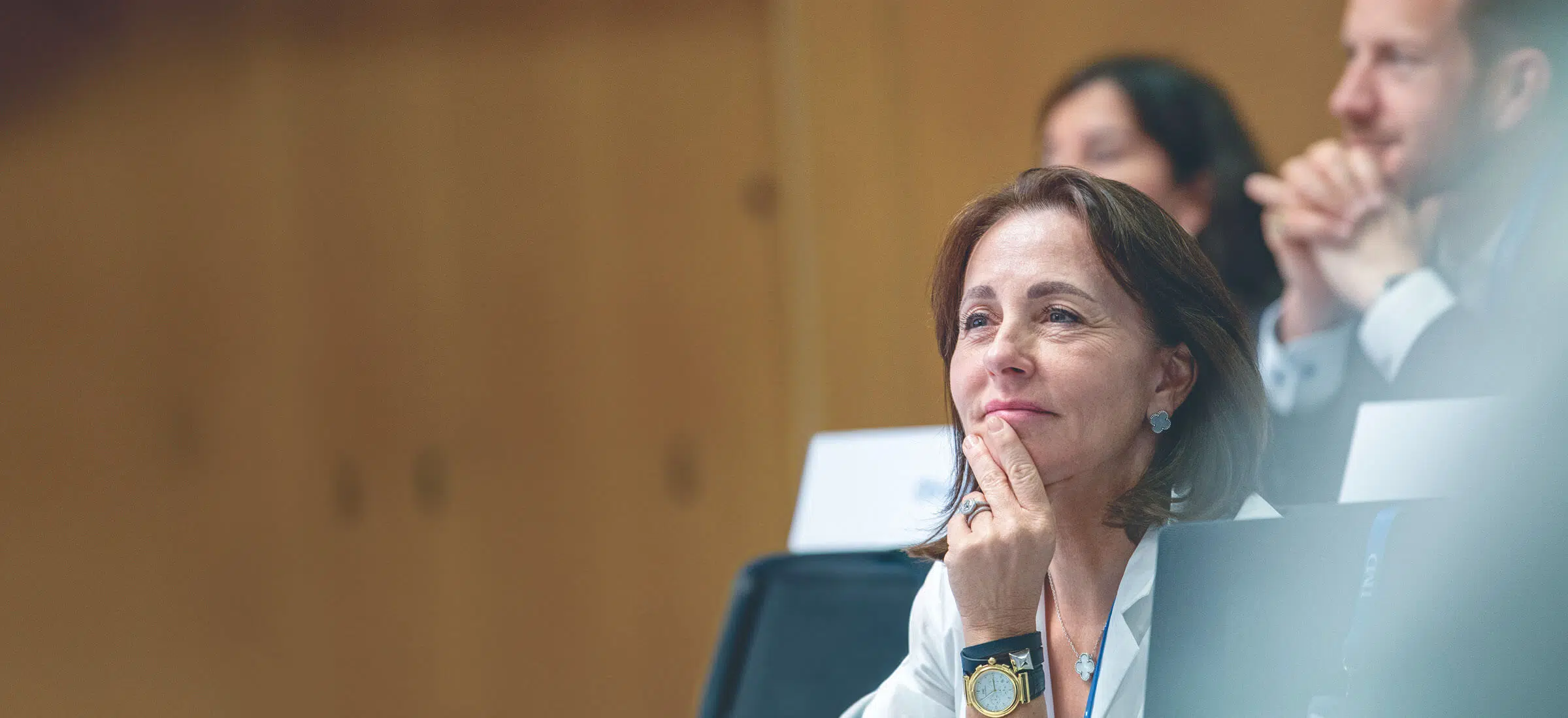 A woman attentively listening during an executive education session, with other participants blurred in the background. - IMD Business School