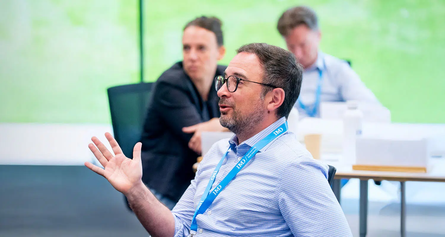 IMD participant speaking during a collaborative workshop session with other attendees in the background. - IMD Business School
