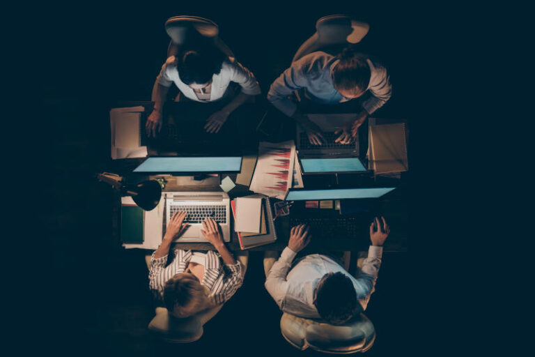 High angle above view photo of four busy partners business men women, working overtime looking computer screen typing corporate email lamp light dark office indoors