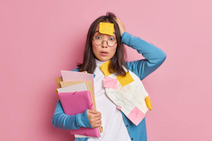 Puzzled shocked female office worker overloaded with paper work stunned to have deadline for finishing research holds folders keeps hand on head wears round spectacles poses against pink background