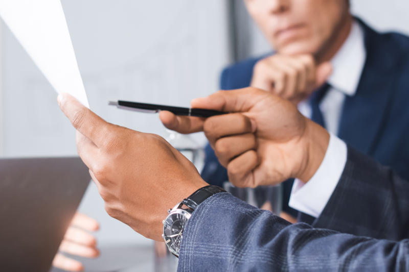 Cropped view of african american businessman pointing with pen at paper with blurred colleagues on background banner