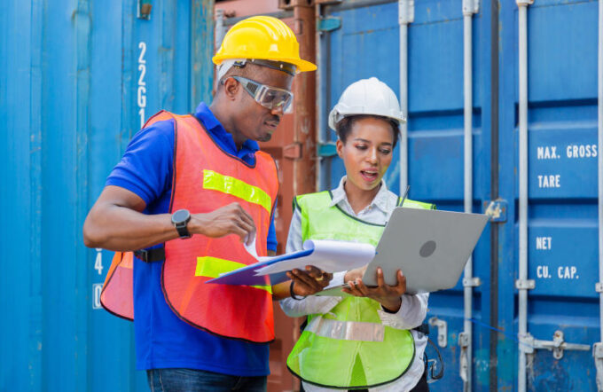 Worker man in hardhat and safety vest holding clipboard checklist and Female foreman using laptop control loading containers box from cargo