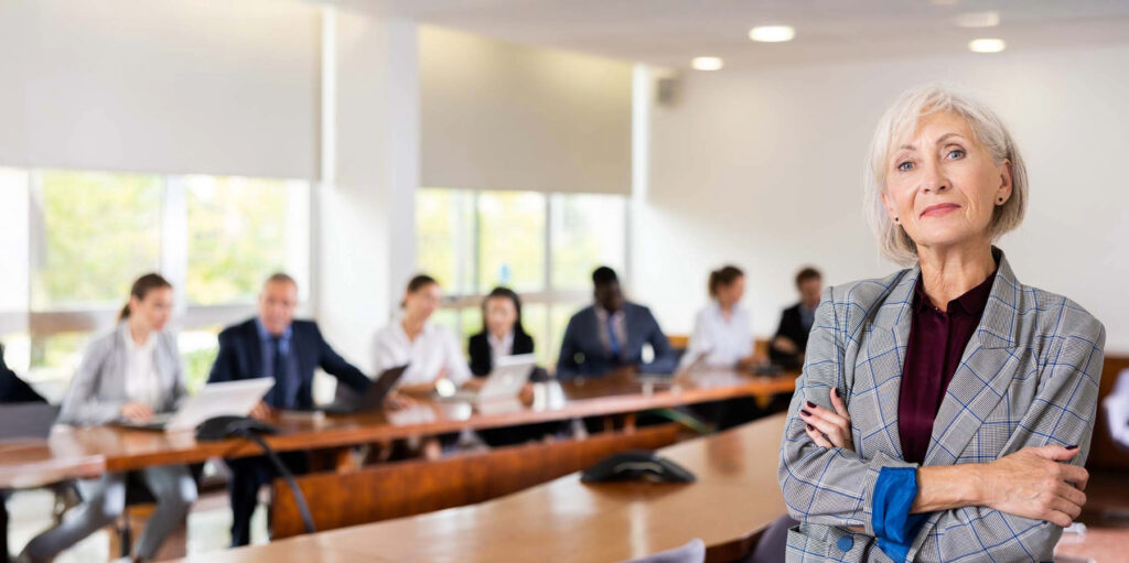 Smiling classy elegant senior mature businesswoman on foreground posing at camera with arms crossed and office employees at back office 2 - IMD Business School Smiling classy elegant senior mature businesswoman on foreground posing at camera with arms crossed and office employees at back office 2 - IMD Business School - IMD Business School