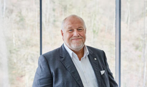 Professional portrait of an older man in a gray suit and white shirt, smiling in a bright, modern office setting.