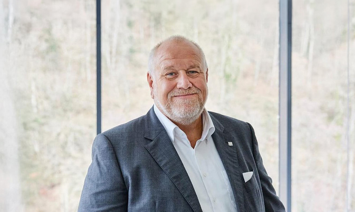 Professional portrait of an older man in a gray suit and white shirt, smiling in a bright, modern office setting.