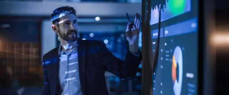 Man in a suit points at a large touchscreen displaying graphs and charts in a high-tech control room. - IMD Business School