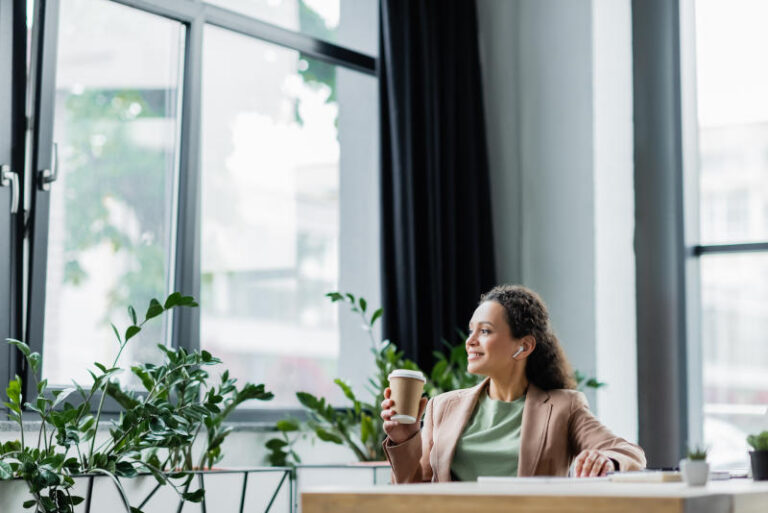 cheerful african american businesswoman listening music in wireless earphone during coffee break in office