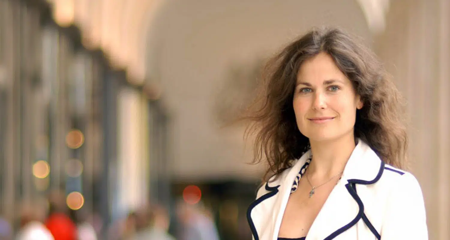 Woman with wavy brown hair in a white blazer with dark trim, smiling outdoors. - IMD Business School