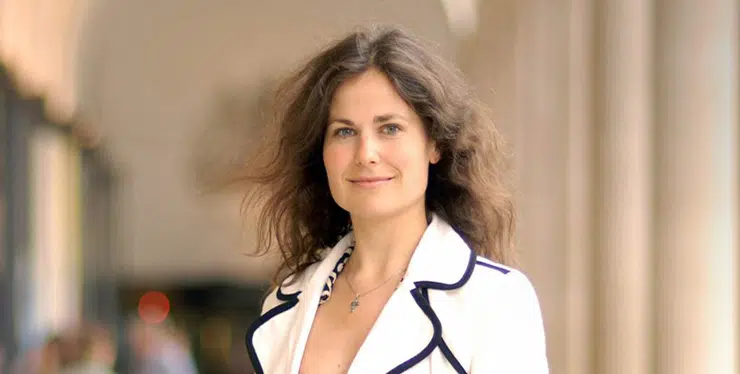Professional-looking woman with shoulder-length brown hair and a white blazer with dark piping, smiling outdoors near a columned walkway. - IMD Business School