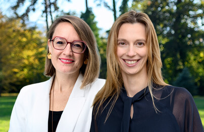 Two smiling women stand together outdoors in a park; one wears a white blazer and red glasses, the other a navy blouse. - IMD Business School