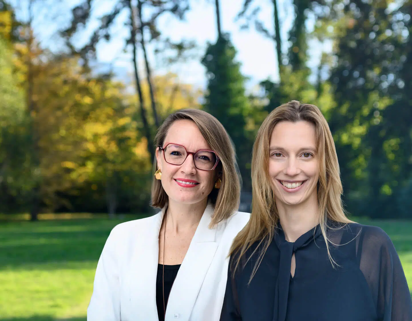 Two women stand together outdoors in a sunlit park, smiling at the camera, one in a white blazer and glasses, the other in a dark blouse. - IMD Business School
