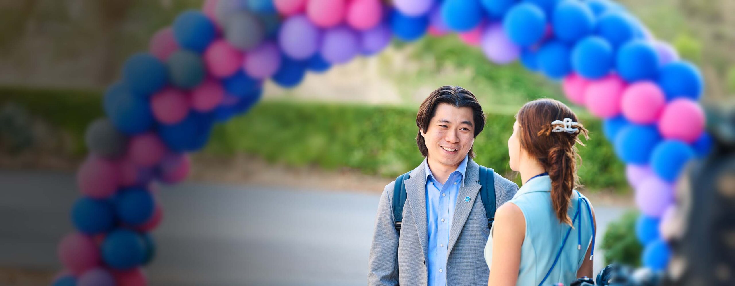 Two people chatting under a colorful balloon arch at an outdoor event; a man in a blazer with a backpack smiles at a woman with braided hair. - IMD Business School