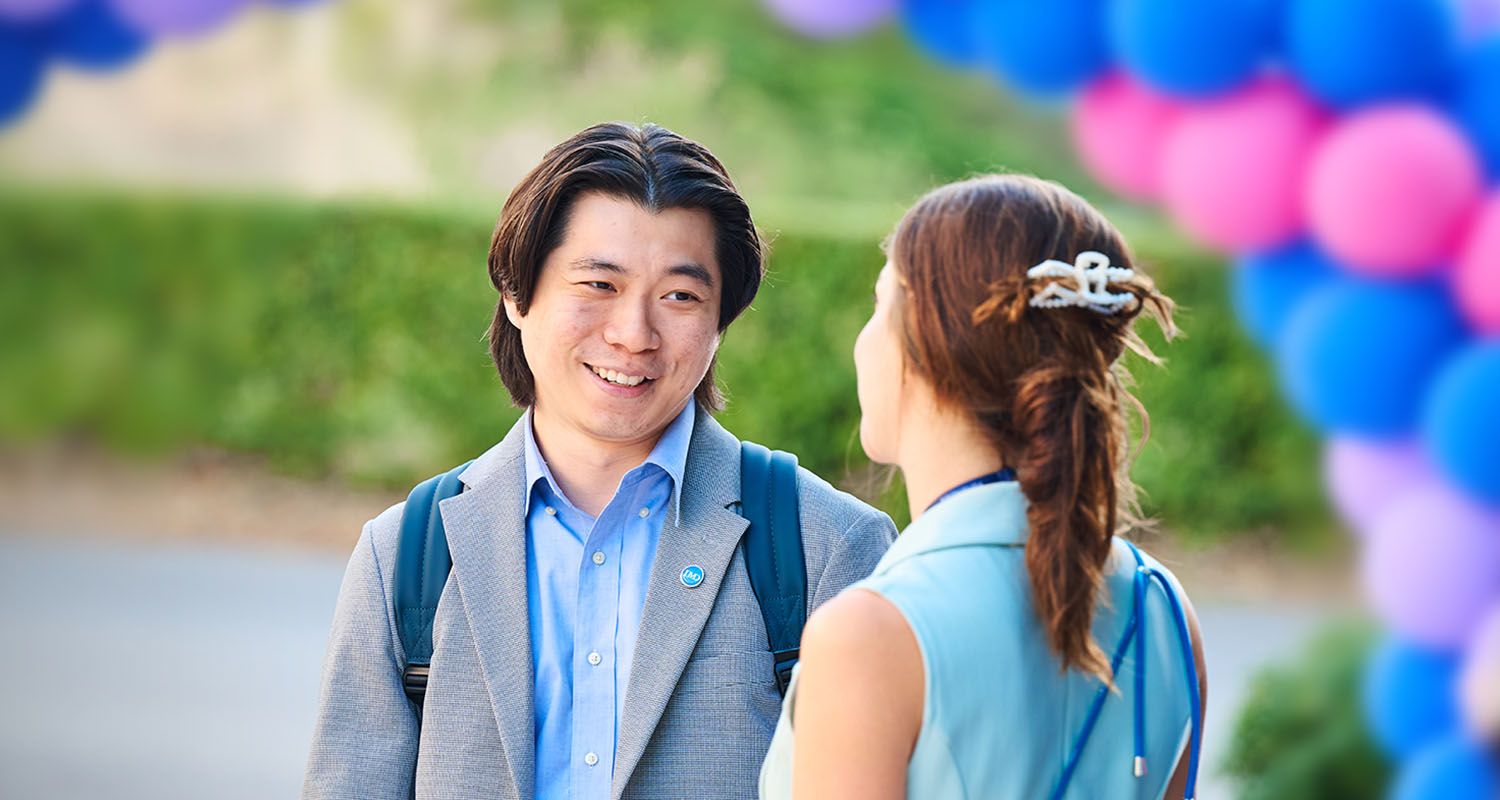 Two people talking outdoors, smiling, beside a colorful balloon arch in the background. - IMD Business School