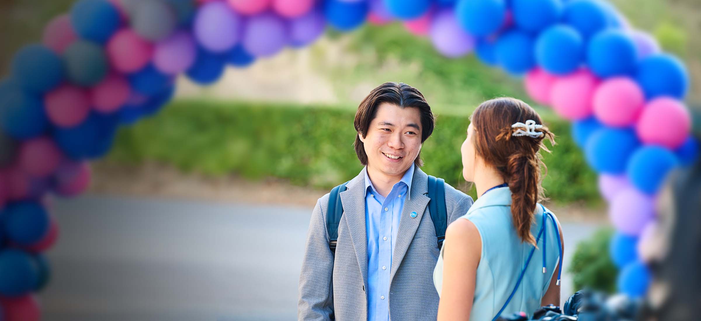 Two people converse under a colorful balloon arch at an outdoor event; man in gray blazer and blue shirt smiles at a woman in a light blue dress with a backpack nearby. - IMD Business School