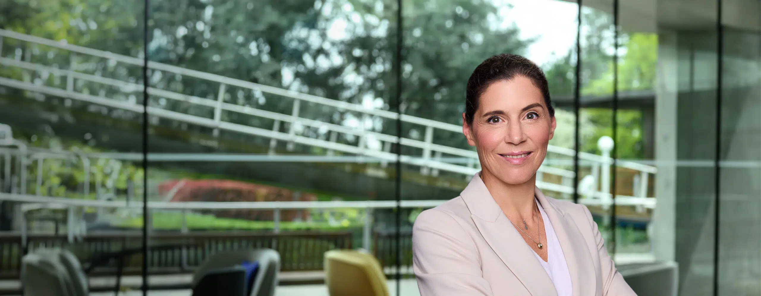 Smiling professional woman with brown hair in a beige blazer stands with arms crossed in a bright glass-walled office setting. - IMD Business School