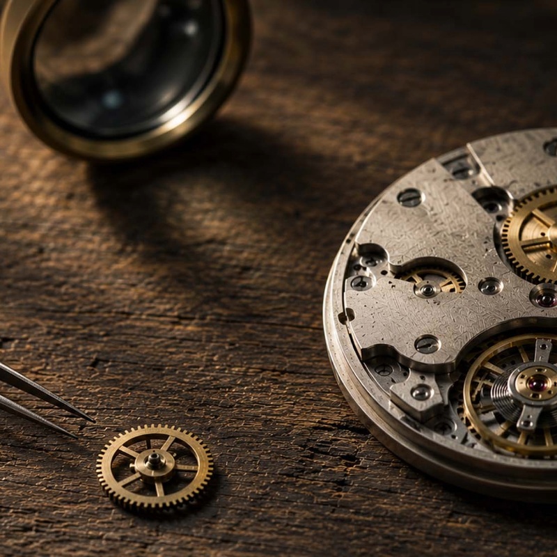 Close-up of a metal watch movement with gears on a wooden workbench, a small gear placed beside tweezers ready for assembly.