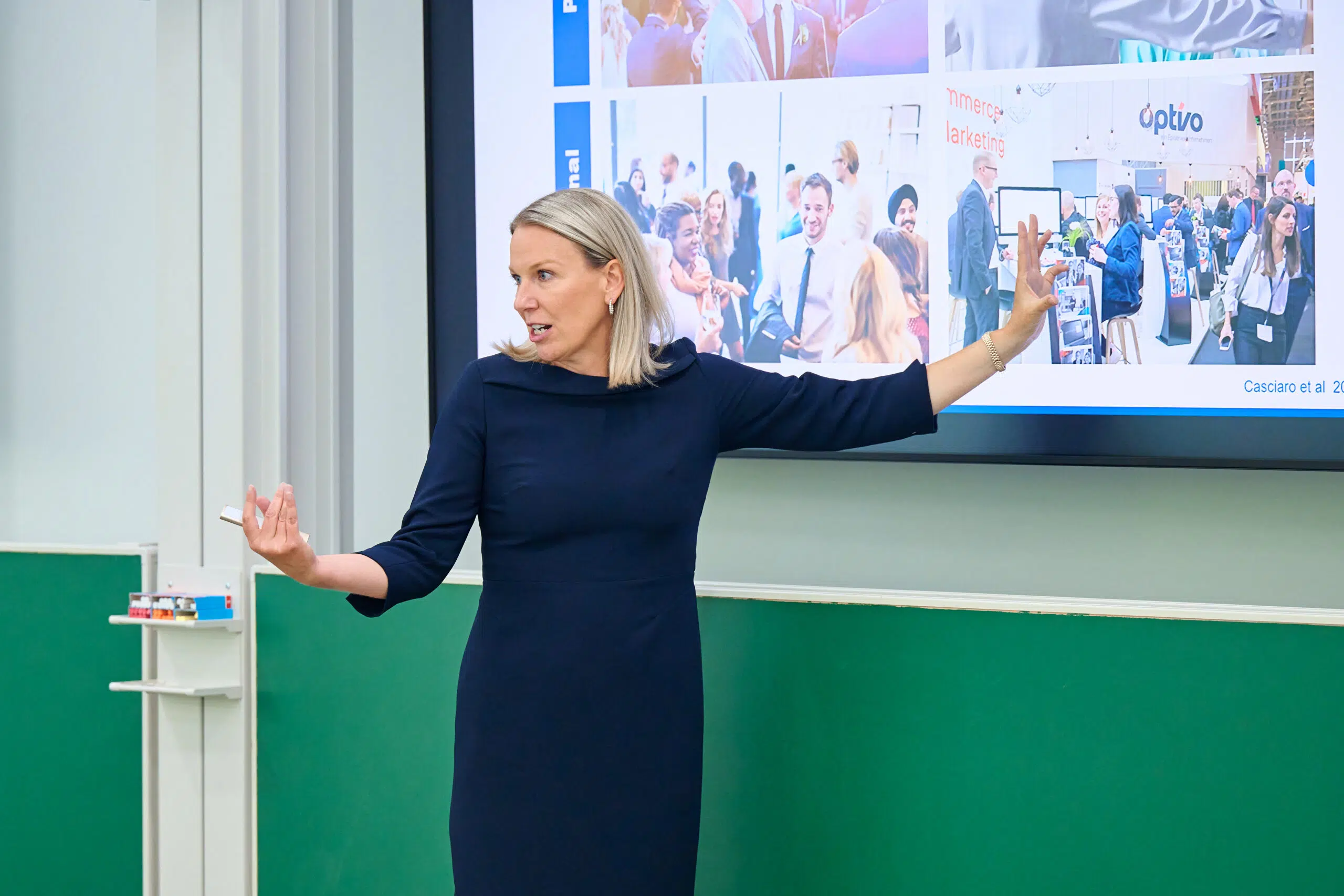 Woman in a navy dress presenting, gesturing with her hands in front of a large screen showing a collage of conference scenes - IMD Business School