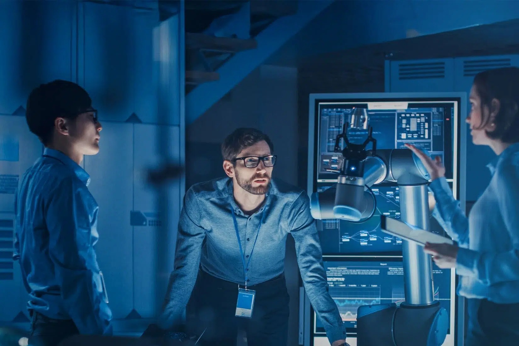 Three researchers in a blue-lit lab study data on a large screen next to an industrial robot arm by a workstation. - IMD Business School
