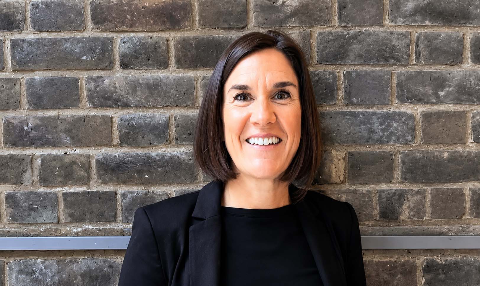Amy Bradley Portrait of a smiling woman with short brown hair wearing a black blazer against a gray brick wall.