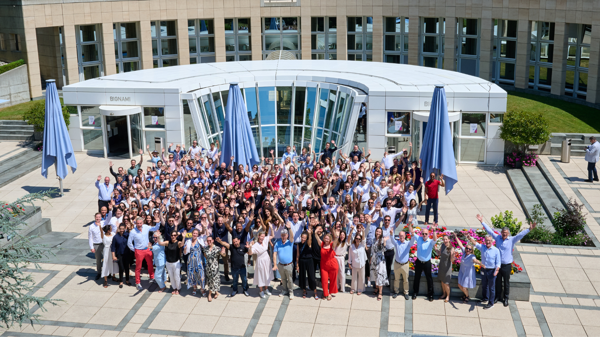 Large group of people waving in front of a modern building with a curved glass entrance. - IMD Business School