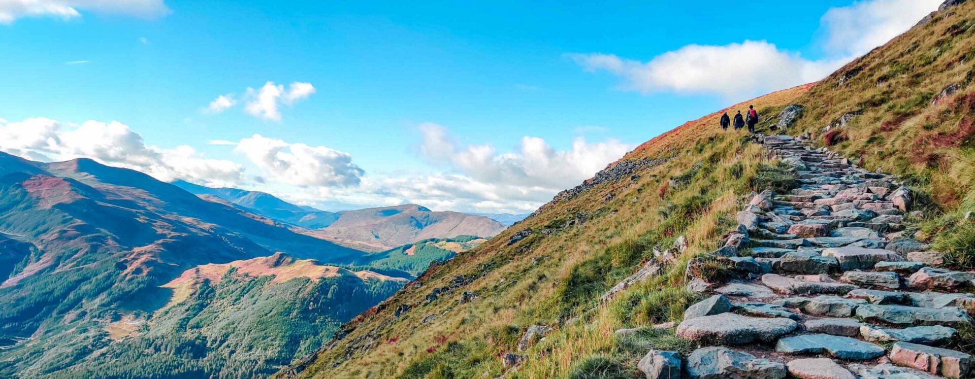 Three hikers ascend a stone staircase along a grassy mountainside.