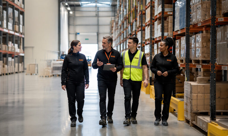 Four warehouse workers in GXO uniforms walk down an aisle of tall shelves with boxes, one holding a tablet.