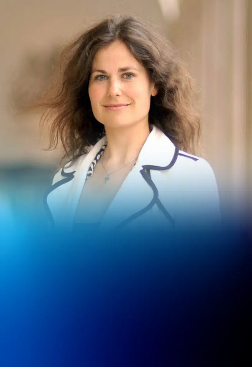 Portrait of a smiling woman with brown, wavy hair wearing a white blazer outdoors, blurred blue foreground - IMD Business School