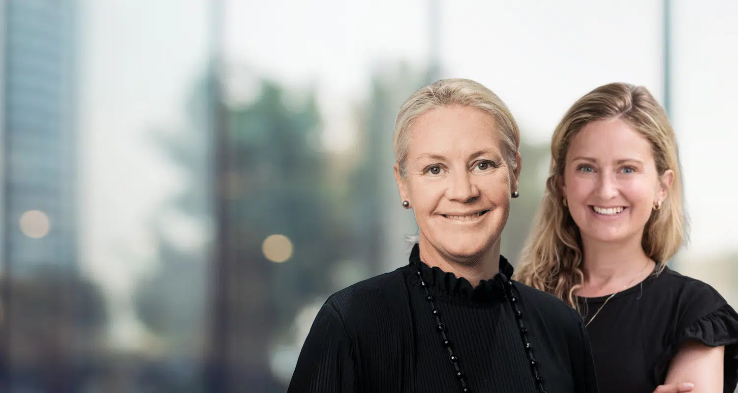 Two smiling professional women pose together in an office, one in front wearing black with a beaded necklace. - IMD Business School