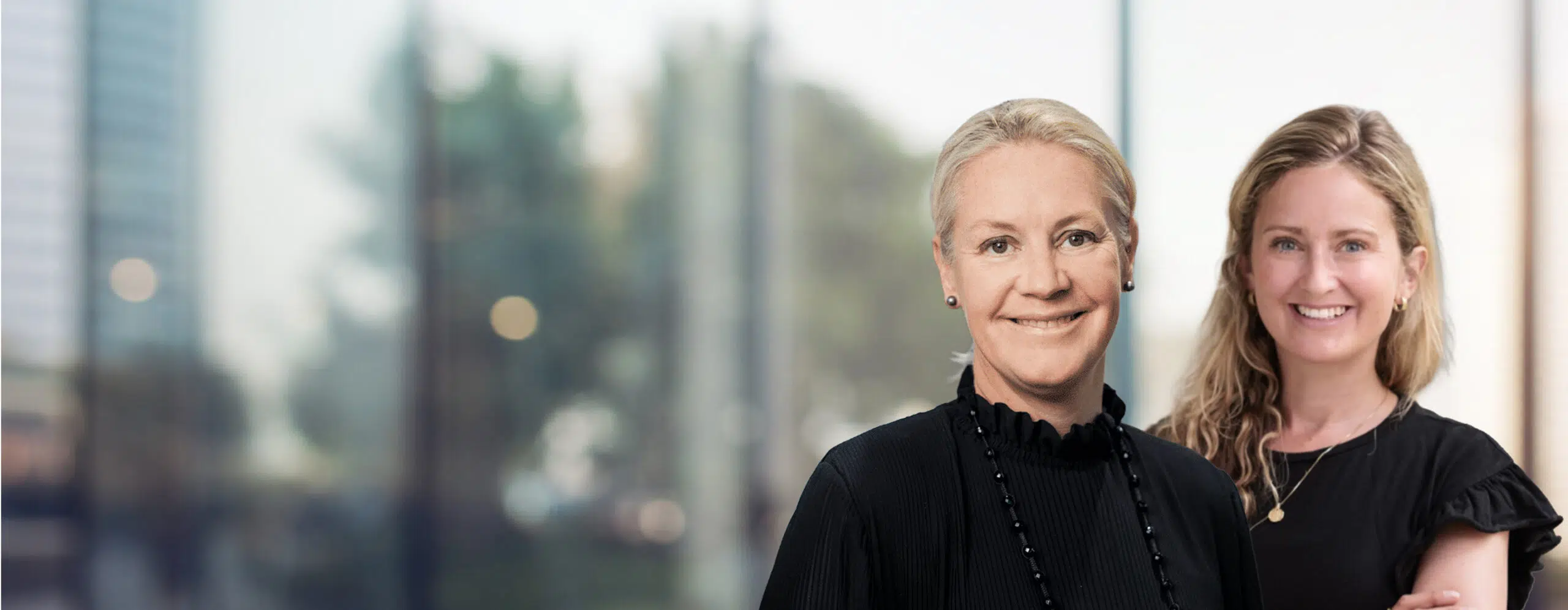 Two smiling women standing together in a bright office setting, both wearing black tops. - IMD Business School