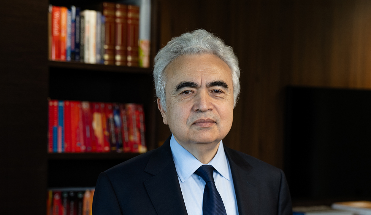 Portrait of an older man with gray hair in a navy suit and blue tie, in a study with bookshelves behind him.