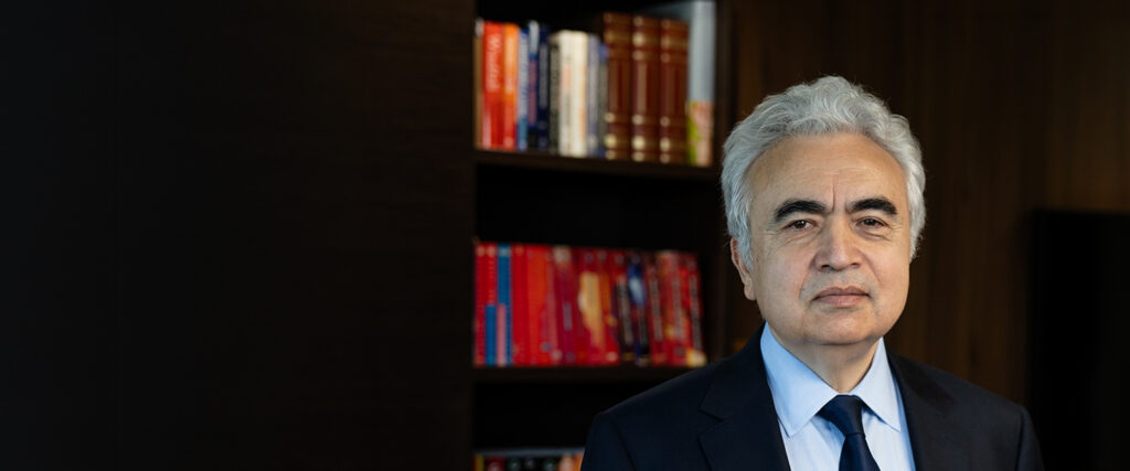 Professional portrait of a middle-aged man with silver hair in a navy suit and blue tie, standing in front of a dark wood bookshelf. - IMD Business School - IMD Business School