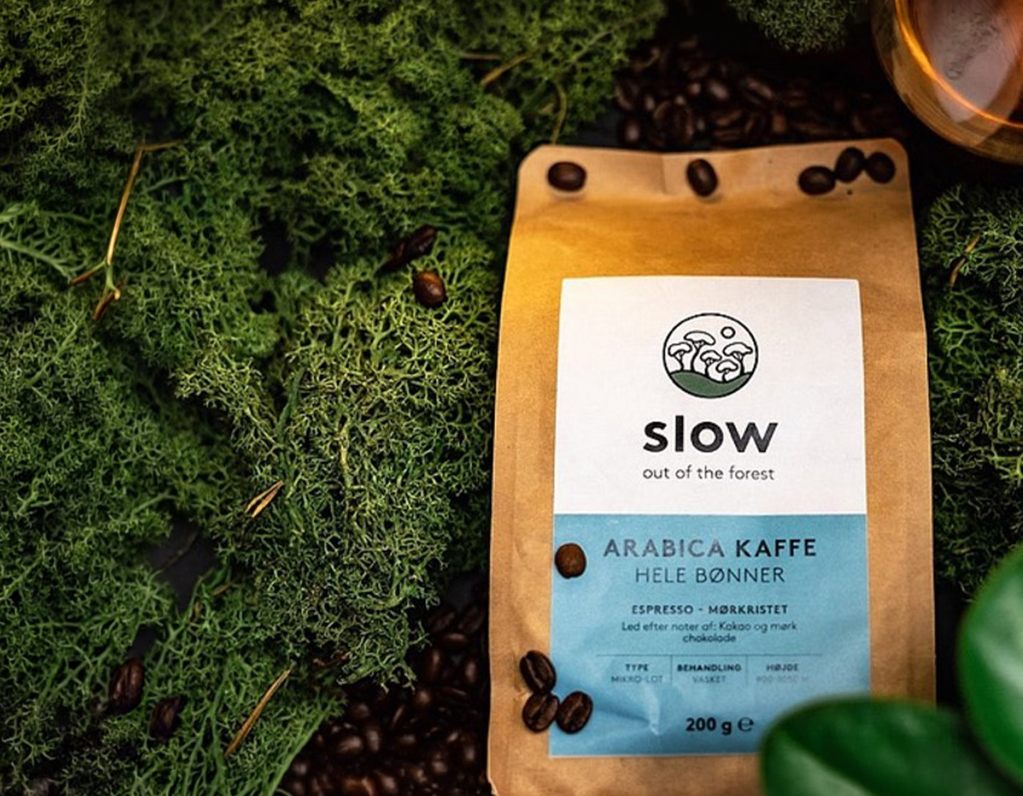 Brown coffee bag on green moss, label reads 'slow out of the forest' and 'Arabica Kaffe Hele Bønner' with scattered roasted beans nearby. - IMD Business School