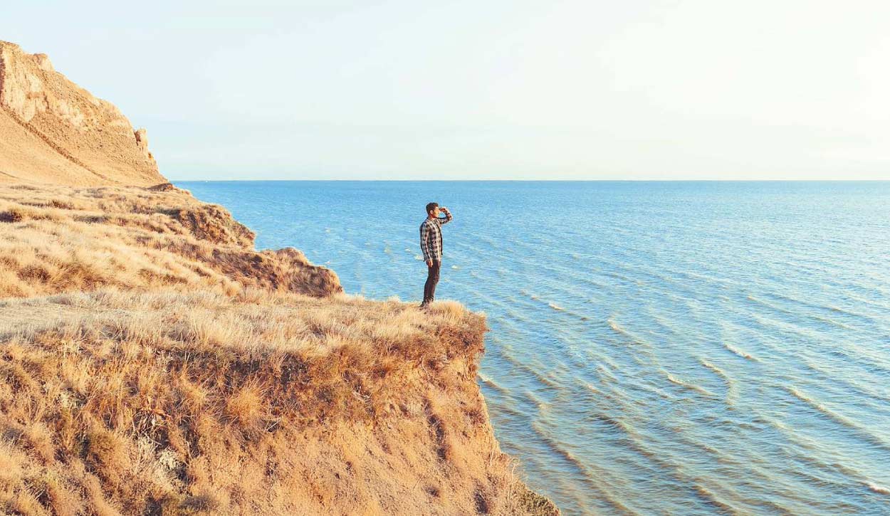Man standing on a grassy cliff edge, looking out over a calm blue sea to the right.