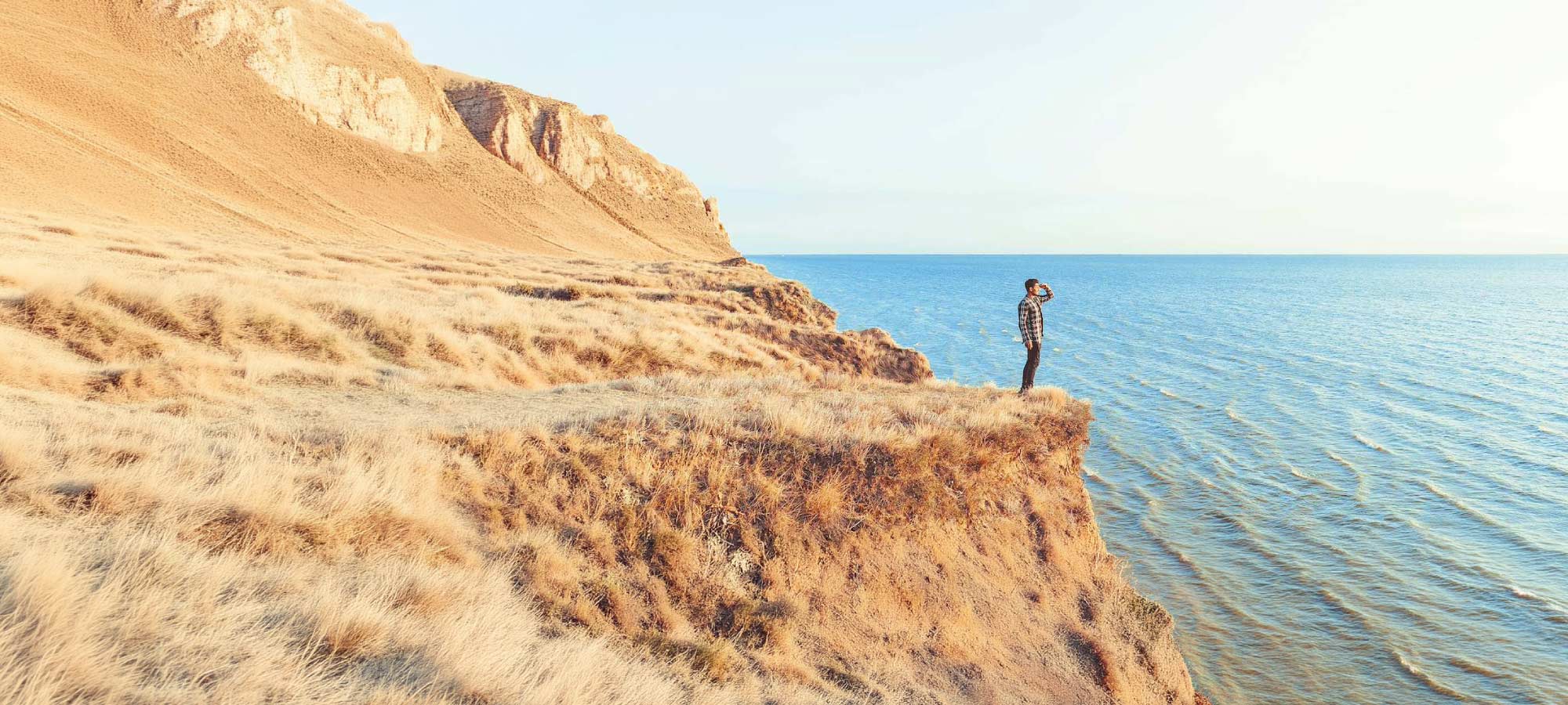 Man standing on a grassy cliff edge, looking out over a calm blue sea to the right.