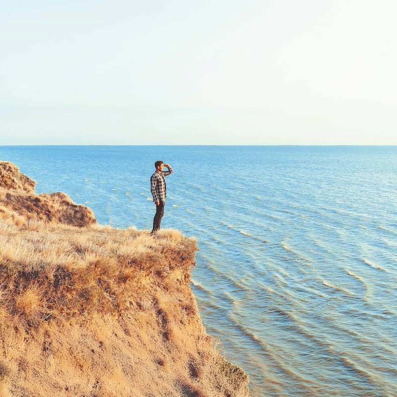Man stands on a rocky cliff at the edge, looking out over a calm blue sea on a sunny day.