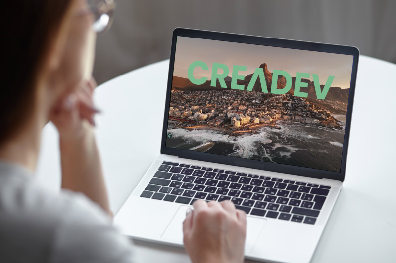 Person sitting at a white table using a laptop; screen shows a coastal city landscape with a turquoise 'CREADEV' logo.
