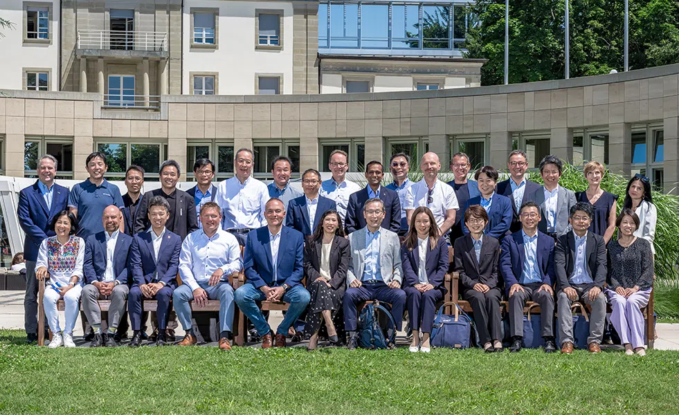 Group portrait of about 30 professionals in business attire posing outdoors in front of a curved building. - IMD Business School