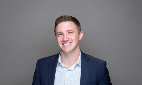 Head-and-shoulders portrait of a smiling man in a navy blazer and a light patterned shirt against a gray background.