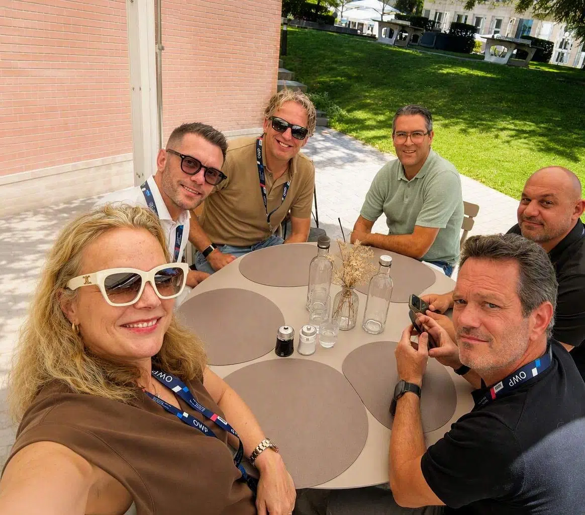 Six people sit around a round outdoor table smiling at the camera, wearing lanyards on a sunny day. - IMD Business School