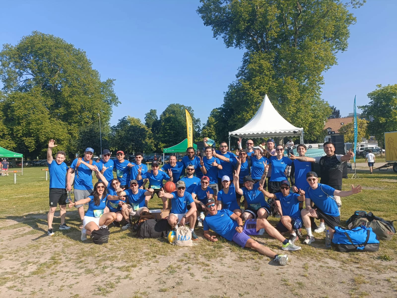 Group of about 30 people in matching blue shirts posing for a photo outdoors at a sunny park with tents in the background. - IMD Business School
