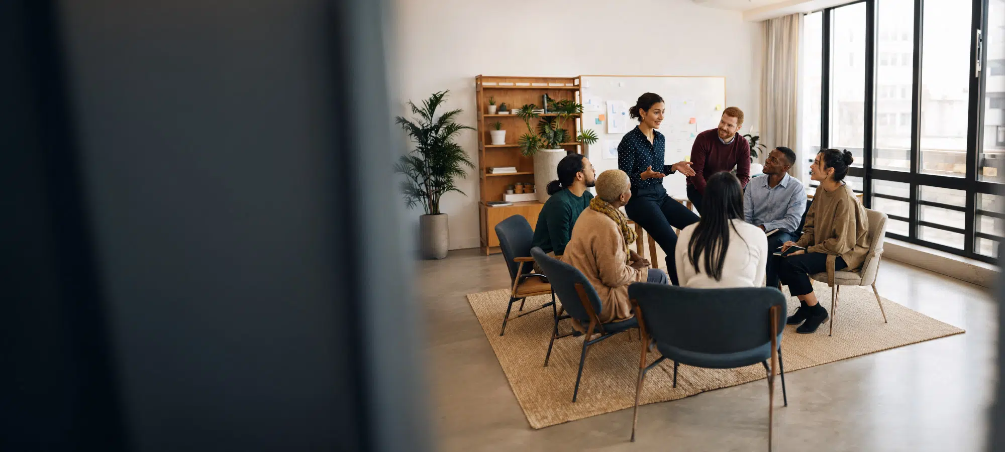 Diverse team in a casual meeting circle in a modern office, one woman standing and presenting while others listen.] - IMD Business School
