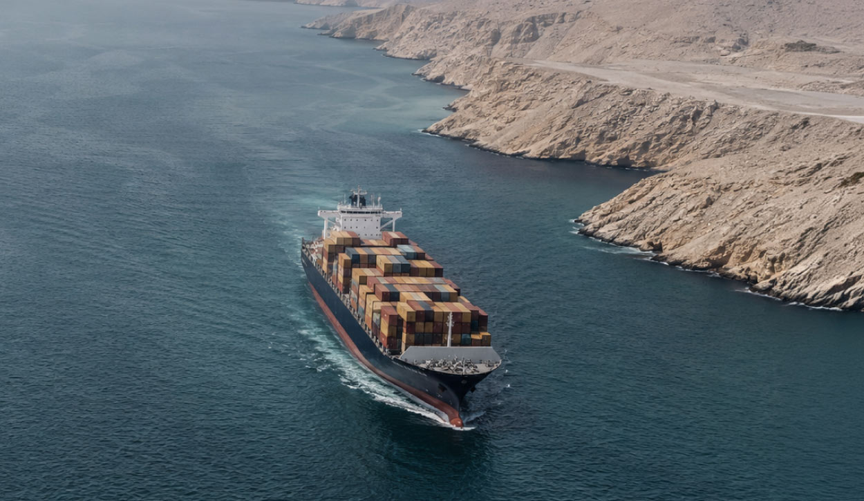 Container ship laden with multicolored shipping containers sails through a narrow waterway between rocky desert cliffs.