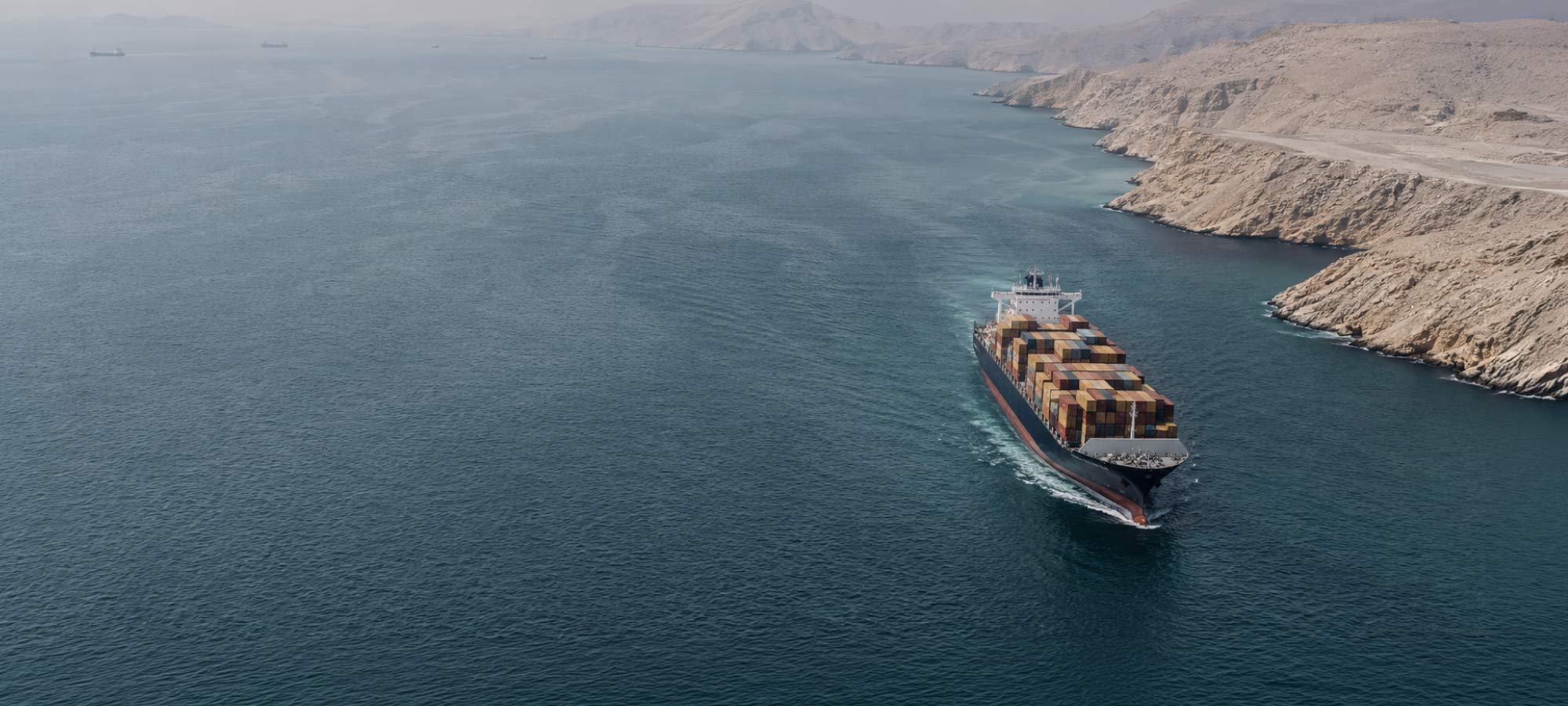 Container ship laden with multicolored shipping containers sails through a narrow waterway between rocky desert cliffs.