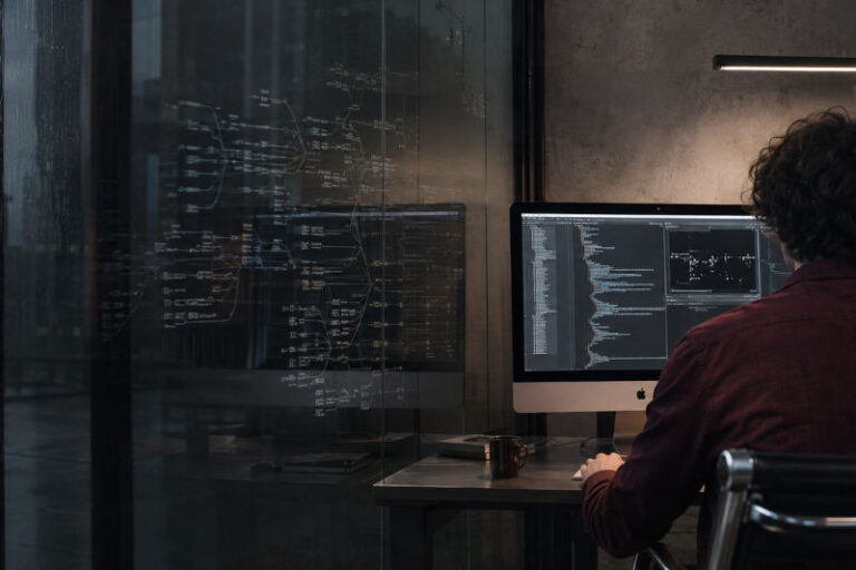 Software developer at a dual-monitor workstation, code on the screens in a dim, modern office.