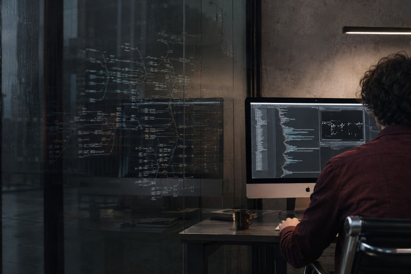 Software developer at a dual-monitor workstation, code on the screens in a dim, modern office.