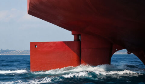 Close-up of the bow of a large red-and-black ship hull looming over the blue sea, with a distant coastline in the background and waves at the stern area.