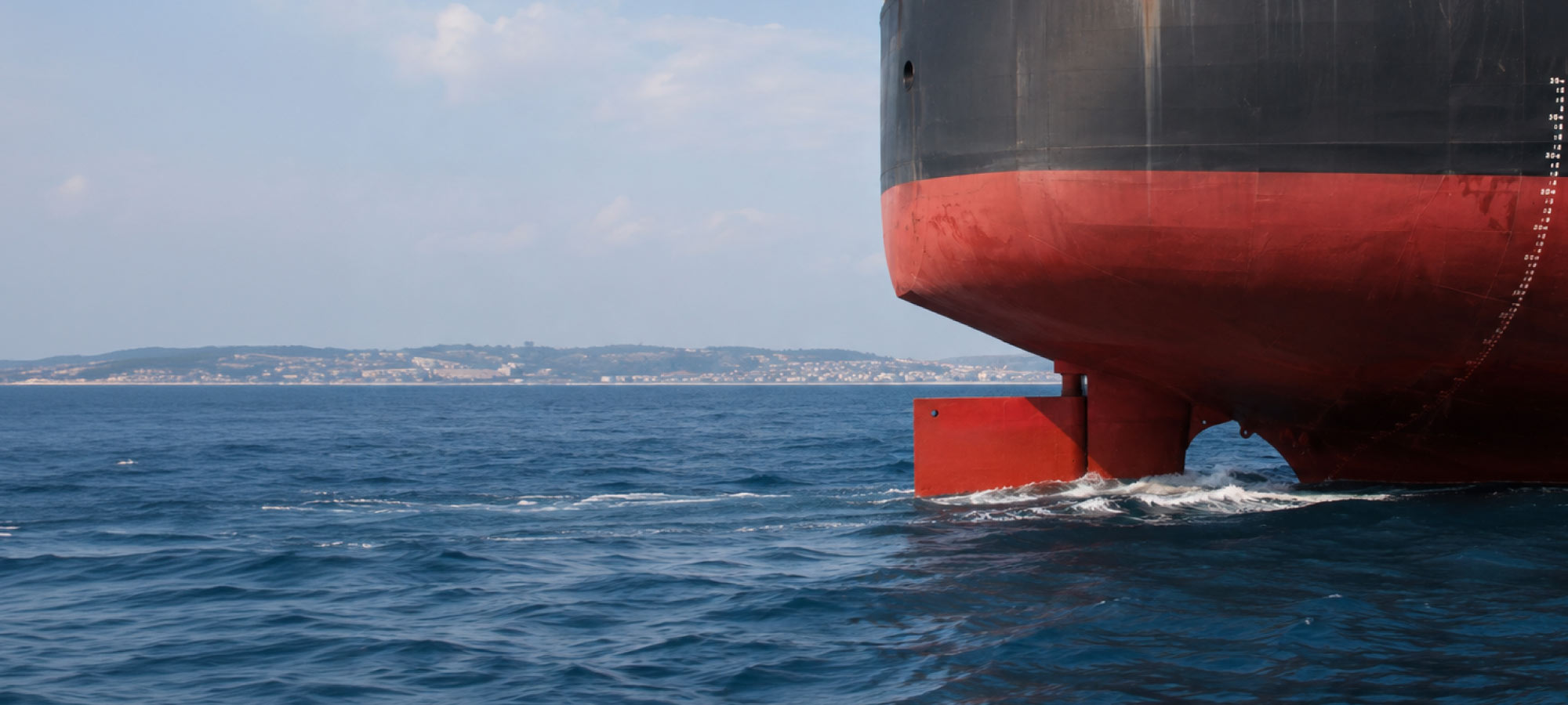 Close-up of the bow of a large red-and-black ship hull looming over the blue sea, with a distant coastline in the background and waves at the stern area.