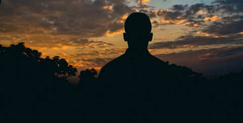 person standing alone beneath a dramatic sunset sky, observing the wider landscape from a position slightly apart, symbolizing independent perspective and insight